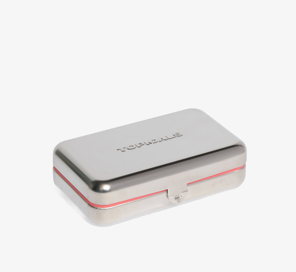 Skincare travel case from Topicals shown as a closed silver metal tin with embossed branding and a pink trim detail on a white background.